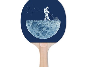May include: A ping pong paddle with a navy blue face featuring an astronaut mowing the moon's surface. The handle is made of light-colored wood. The design includes stars and a detailed depiction of the moon.