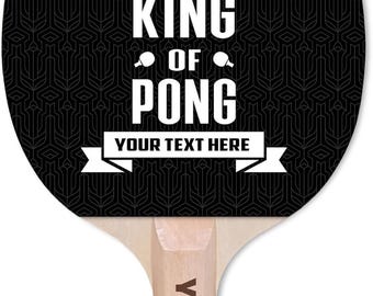 May include: A black and white ping pong paddle with the text "KING OF PONG" and "YOUR TEXT HERE". The paddle has a wooden handle and a black patterned face with a white crown graphic.