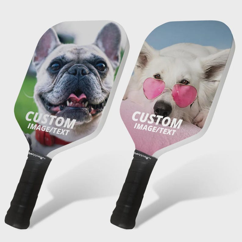 May include: Two white pickleball paddles with custom images of dogs. The paddle on the left has a French Bulldog with the text "CUSTOM IMAGE/TEXT". The paddle on the right has a white dog wearing pink heart-shaped sunglasses with the text "CUSTOM IMAGE/TEXT".