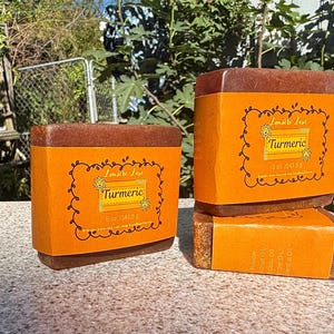 Puede incluir: Tres pastillas de jabón de cúrcuma rectangulares envueltas en etiquetas de papel naranja. Cada etiqueta tiene un borde negro decorativo, el texto "Lumière Luxe", "Turmeric" y "5 oz / 141,5 g". Los jabones tienen un aspecto rojizo oscuro y texturizado.