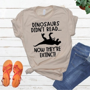 May include: A beige t-shirt with a black graphic that says "Dinosaurs didn't read... Now they're extinct!" and a silhouette of a dinosaur falling on its back.