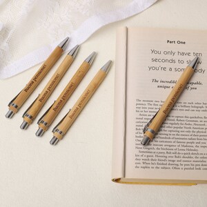 May include: Four brown bamboo pens with silver accents and the text "Buona pensione!" engraved on each pen. The pens are lying on a white surface next to an open book.