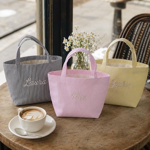 May include: Three small tote bags with two handles each, featuring vertical pinstripes in black, pink, and yellow. Each bag has a name embroidered in white script on the front: Laura on the black bag, Mia on the pink bag, and Sophie on the yellow bag.