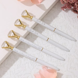 Könnte beinhalten: Vier weiße Stifte mit goldenen Akzenten und klaren Strasssteinen. Die Stifte sind mit dem Text "Bride Tribe" und "Matter & Chase 15 August 2024" personalisiert.