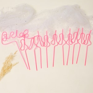 May include: Pink plastic straws shaped like the word "bride" and other designs. The straws are on a white background with a white sparkly fabric in the background.