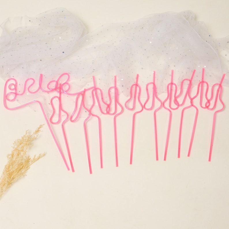 May include: Pink plastic straws shaped like the word "bride" and other designs. The straws are on a white background with a white sparkly fabric in the background.