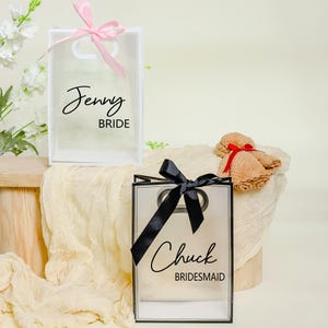 May include: Two clear plastic gift bags with black and white text. The bag on the left has a pink ribbon and says "Jenny Bride". The bag on the right has a black ribbon and says "Chuck Bridesmaid".