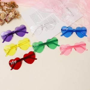 Puede incluir: Siete pares de gafas de sol en forma de coraz&oacute;n en varios colores, incluyendo rojo, amarillo, verde, azul, morado y rosa. Cada par tiene un nombre diferente escrito en blanco en las lentes.