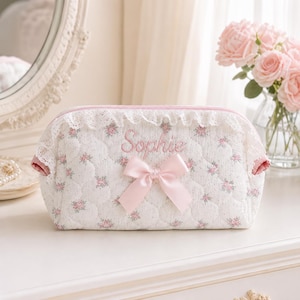 May include: A quilted white cosmetic bag featuring a small pink floral pattern, a pink embroidered name Sophie, a pink satin ribbon bow, and white lace trim along the top edge. The bag has a zip closure and pink fabric lining on the sides.