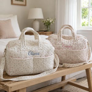 May include: Two quilted duffel bags with a small floral pattern. The bag on the left is white with blue flowers and the name Olivia embroidered in blue. The bag on the right is a soft pink with pink flowers and the name Evie embroidered in pink.