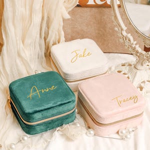Könnte beinhalten: Drei Schmuckkästchen aus Samt mit goldenen Reißverschlüssen. Die Kästchen sind in Grün, Rosa und Weiß. Die Kästchen sind mit den Namen "Anne", "Tracey" und "Jake" personalisiert.