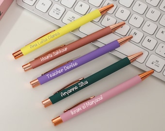 May include: Five personalized pens with rose gold accents. The pens are yellow, orange, purple, green, and pink. The names on the pens are Ana Luisa Clares, Houria Sakkour, Teacher Denise, Aryanne Silva, and Ikram el Maryaoui.