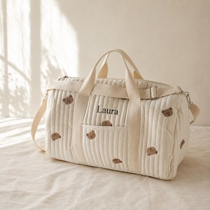 May include: A cream-coloured quilted duffel bag featuring an embroidered pattern of small brown teddy bears. The bag includes two top handles, a detachable shoulder strap with metal hardware, and the name Laura embroidered in black on the front.