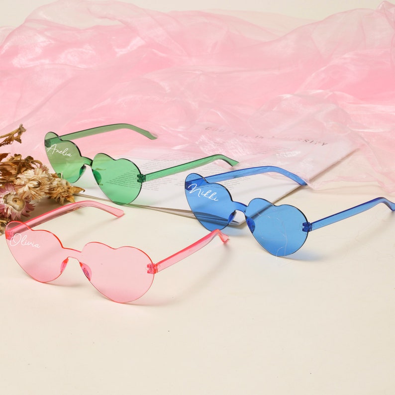 Puede incluir: Tres pares de gafas de sol con forma de coraz&oacute;n con diferentes colores de montura y lentes. Las gafas de sol est&aacute;n personalizadas con los nombres Olivia, Milla y Andia.