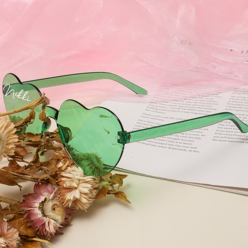 Puede incluir: Gafas de sol verdes en forma de coraz&oacute;n con el nombre "Nikki" escrito en la lente izquierda. Las gafas de sol est&aacute;n apoyadas en un libro y un ramo de flores secas.