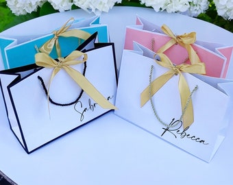 May include: Three white gift bags with black trim and gold ribbon. The bags are personalized with the names "Sabrina", "Rebecca", and a third name that is not visible in the image.