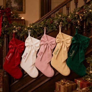 May include: Five velvet Christmas stockings in red, white, pink, gold, and dark green, each featuring a large matching bow and embroidered names: Emma, Olivia, Lily, Grace, and Noah. The stockings are hung in a row, each with a loop for display.