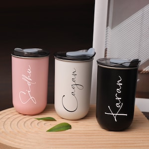 May include: Three personalized stainless steel travel mugs with lids. The mugs are pink, white, and black. The names "Siddh", "Cagjan", and "Karaan" are written in cursive on the mugs.