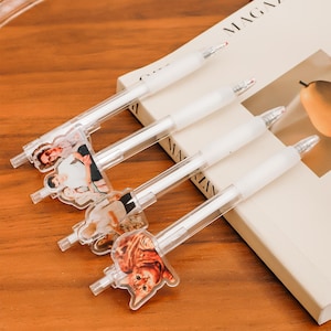 Op de afbeelding: Vier transparante plastic pennen met verschillende afbeeldingen aan de uiteinden. De afbeeldingen omvatten een ​​familiefoto, een kat en twee andere foto's. De pennen liggen op een boek met het woord "MAGAZINE" zichtbaar op de omslag.