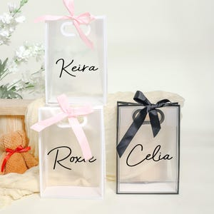 May include: Three clear gift bags with personalized names. The bags are decorated with pink and black ribbon bows. The names on the bags are Keira, Roxie, and Celia.