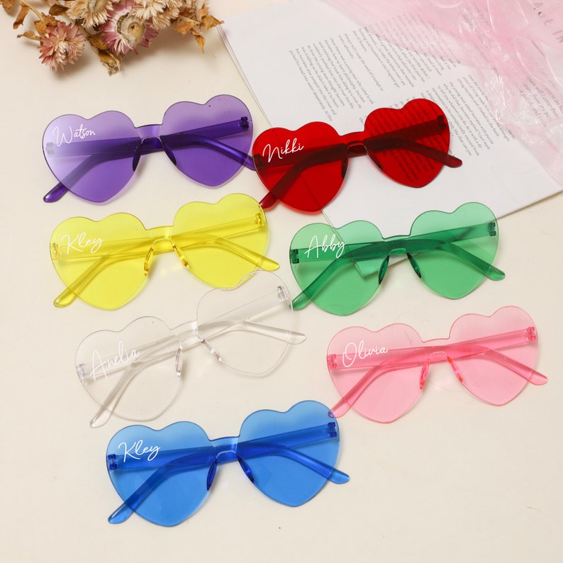 Puede incluir: Conjunto de ocho gafas de sol en forma de coraz&oacute;n de varios colores, cada una con un nombre diferente escrito en la lente. Las gafas de sol est&aacute;n dispuestas en semic&iacute;rculo sobre una superficie blanca.