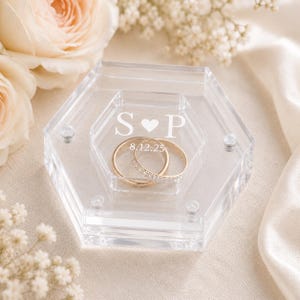 May include: A clear hexagonal acrylic ring box featuring the engraved letters S and P separated by a heart symbol, with the date 12.08.25 below. Two gold-toned metal rings, one with a pavé-set band, are resting inside the transparent container.
