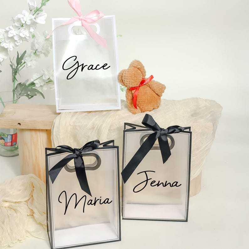 May include: Three clear gift bags with black and white trim. The bags have black ribbon bows and are personalized with the names "Grace", "Jenna", and "Maria".