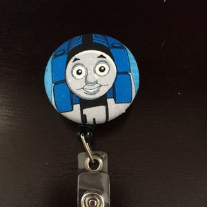 May include: A blue and white Thomas the Tank Engine badge reel. The reel features a cartoon image of Thomas's face. The reel has a clear plastic clip and a black cord.