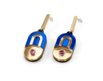 Peut inclure: Une paire de boucles d'oreilles géométriques. Chaque boucle d'oreille présente une tige rectangulaire dorée, reliée à un pendentif bleu, doré et noir. Une petite gemme rose est sertie au centre de la base dorée. Les boucles d'oreilles sont sur fond blanc.