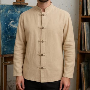 May include: A beige long-sleeved shirt with a mandarin collar and wooden button closures. The shirt is made of a light-colored fabric and is buttoned up the front. The shirt is part of a clothing collection.