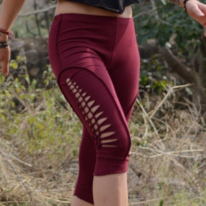 Peut inclure: Un legging capri bordeaux avec des détails découpés sur les côtés. Le legging arrive au genou et est fait d'une matière extensible. La personne porte des sandales et marche sur un sentier de terre.