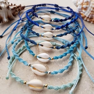 Puede incluir: Un conjunto de seis pulseras de amistad azules y turquesas, cada una con un dije de concha de cauri. Las pulseras están hechas con cordón trenzado y tienen cuentas doradas y plateadas.