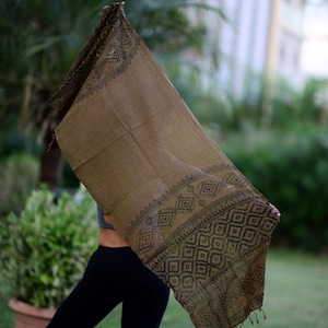 May include: A rectangular brown scarf featuring a dark brown printed geometric pattern along the borders. The fabric has a visible woven texture and includes small fringe details at the bottom edge, showcasing a repeating diamond and square motif.