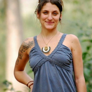 Pu&ograve; includere: Una donna con capelli castano scuro indossa un abito blu senza maniche con una scollatura intrecciata. Indossa una collana d'oro con un ciondolo decorativo e un braccialetto di pelle marrone al polso. Ha un tatuaggio sulla spalla sinistra.