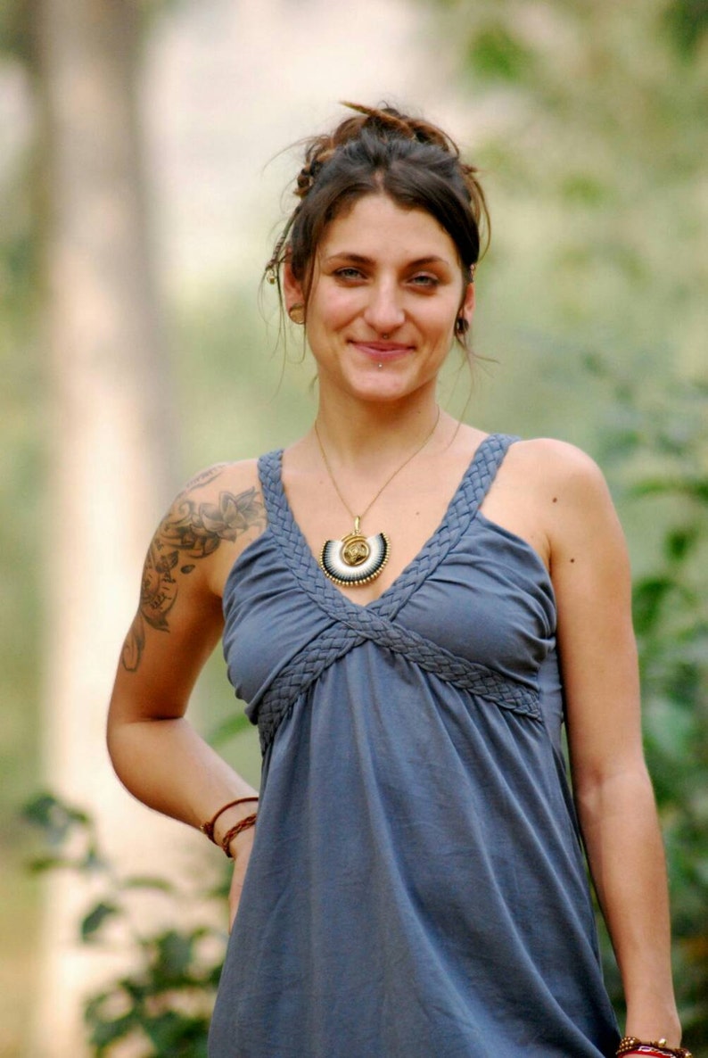 Pu&ograve; includere: Una donna con capelli castano scuro indossa un abito blu senza maniche con una scollatura intrecciata. Indossa una collana d'oro con un ciondolo decorativo e un braccialetto di pelle marrone al polso. Ha un tatuaggio sulla spalla sinistra.