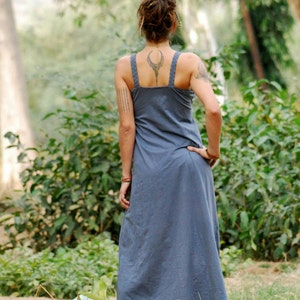 Pu&ograve; includere: Una donna che indossa un lungo abito blu senza maniche con un tatuaggio geometrico sulla schiena. &Egrave; in piedi in un ambiente boschivo.