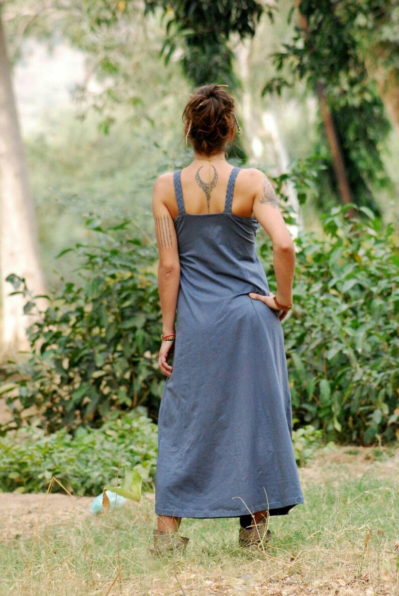 Pu&ograve; includere: Una donna che indossa un lungo abito blu senza maniche con un tatuaggio geometrico sulla schiena. &Egrave; in piedi in un ambiente boschivo.