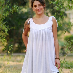 May include: A woman wearing a white sleeveless dress with a gathered bodice and black leggings. She is standing in a grassy area with trees in the background.