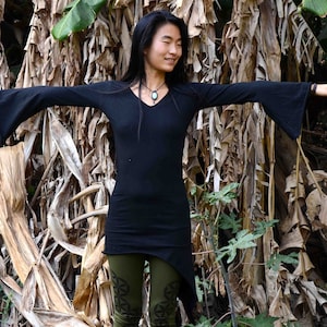 May include: A woman wearing a black long-sleeved shirt with a V-neck and a long, asymmetrical hem. She is standing in front of a background of dried banana leaves.