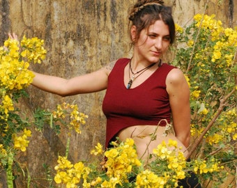 May include: A woman wearing a red cropped top with a beaded necklace stands in front of a yellow flowering bush. The woman has long brown hair and is wearing black pants.