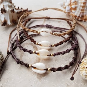 Puede incluir: Cuatro pulseras de cordón trenzado marrón y beige con abalorios de concha de cauri. Las pulseras están superpuestas sobre una superficie blanca.