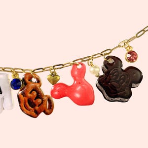 May include: A gold chain necklace featuring a variety of charms. Charms include a blue gem, a brown Mickey Mouse-shaped charm, a red charm, a gold heart, a pearl, a chocolate ice cream sandwich charm, and a pink gem. The charms are suspended from a gold chain.