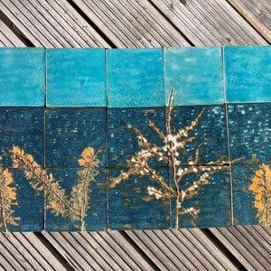 Puede incluir: Un conjunto de 12 azulejos de cerámica con un esmalte azul y verde. Los azulejos presentan un diseño floral con flores blancas y amarillas. Los azulejos están dispuestos en una cuadrícula.