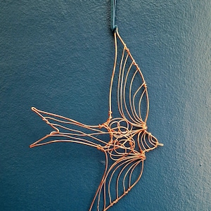 May include: A copper wire sculpture shaped like a bird in flight, featuring intricate wire loops and coils forming the body and wings. A dark blue ribbon is attached to the top for hanging, and a small blue bead hangs from the bottom tail section.