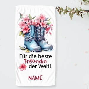 Op de afbeelding: Witte handdoek met een aquarel illustratie van lichtblauwe laarzen gevuld met roze bloemen. De tekst "Für die beste Freundin der Welt!" en "NAME" zijn in zwart en rood gedrukt.