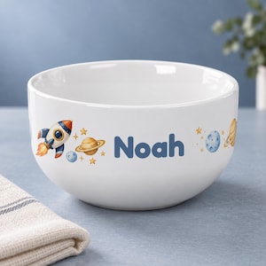 Könnte beinhalten: Eine weiße Keramikschale mit einer bunten Weltraum-Illustration aus einer Rakete, Planeten und Sternen, wobei der Name Noah in fetten blauen Buchstaben an der Seite aufgedruckt ist. Die Schale hat eine glatte, abgerundete Form und Oberfläche.