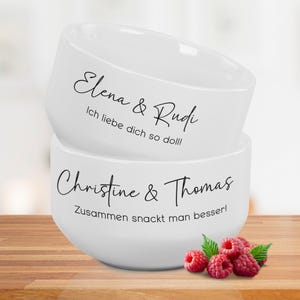 Könnte beinhalten: Zwei weiße Keramikschalen, die übereinander gestapelt sind, jede mit schwarzer Schrift. Auf der oberen Schale steht "Elena & Rudi" und "Ich liebe dich so doll!". Auf der unteren Schale steht "Christine & Thomas" und "Zusammen snackt man besser!". Rote Himbeeren unten.