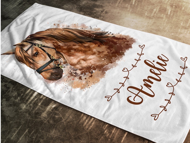 May include: A white beach towel with a watercolor painting of a brown horse with a bridle. The horse's head is turned to the side and its eyes are closed. The name "Amelie" is written in brown cursive script on the towel.