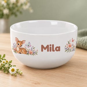 Könnte beinhalten: Eine weiße Keramikschale mit einer aufgedruckten Illustration eines braunen Rehkitzes, das zwischen bunten Blumen und einem Schmetterling ruht. Der Name "Mila" ist in braunen Buchstaben seitlich aufgedruckt, ergänzt durch ein Blumenarrangement.