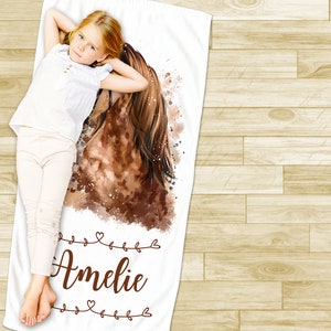 May include: White beach towel with a watercolor painting of a brown horse's head and a girl lying on the towel. The towel has the name "Amelie" written in brown cursive with decorative hearts.